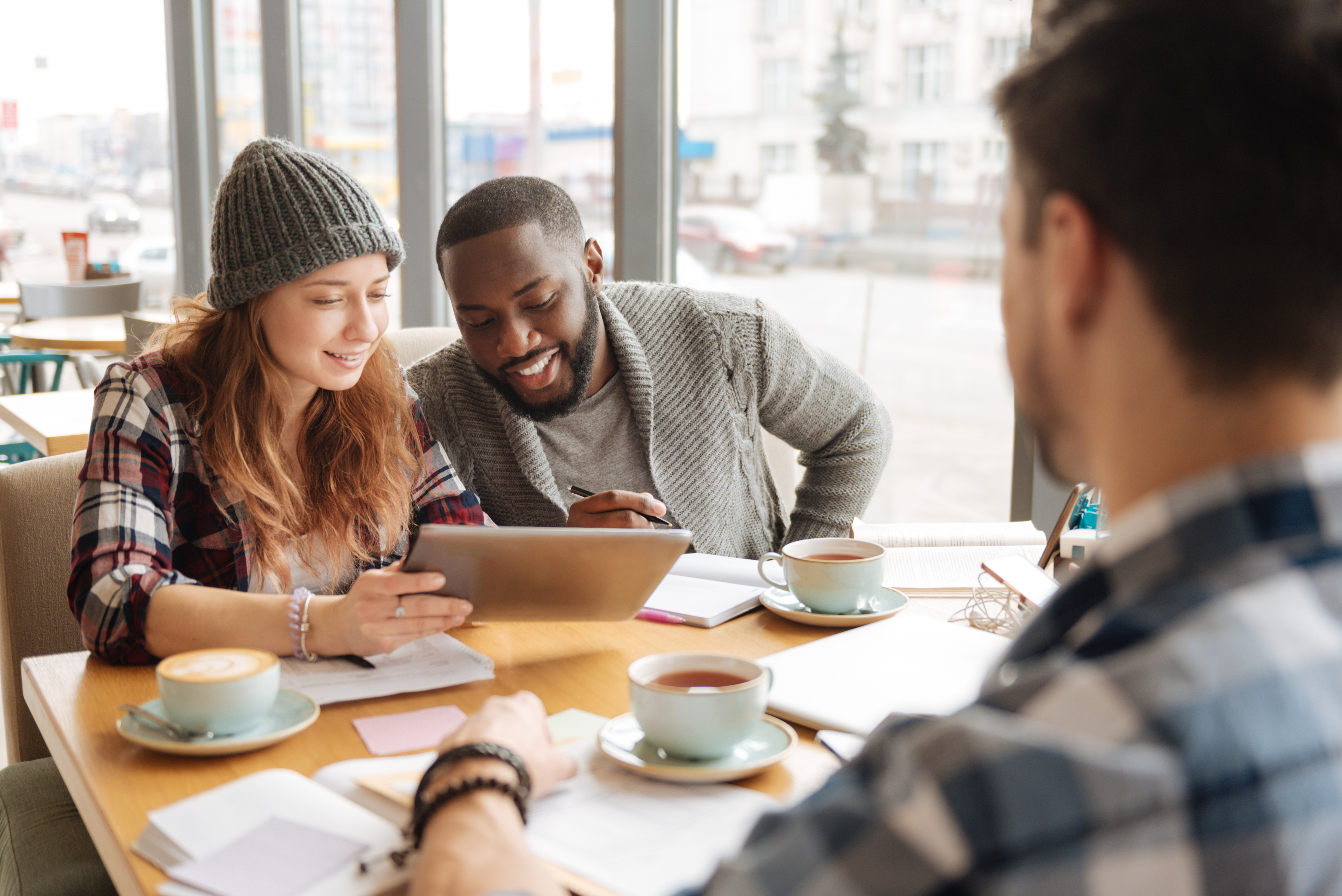 En grupp människor sitter på ett café och dricker kaffe och tittar tillsammans på en surfplatta, papper och anteckningsböcker ligger på bordet
