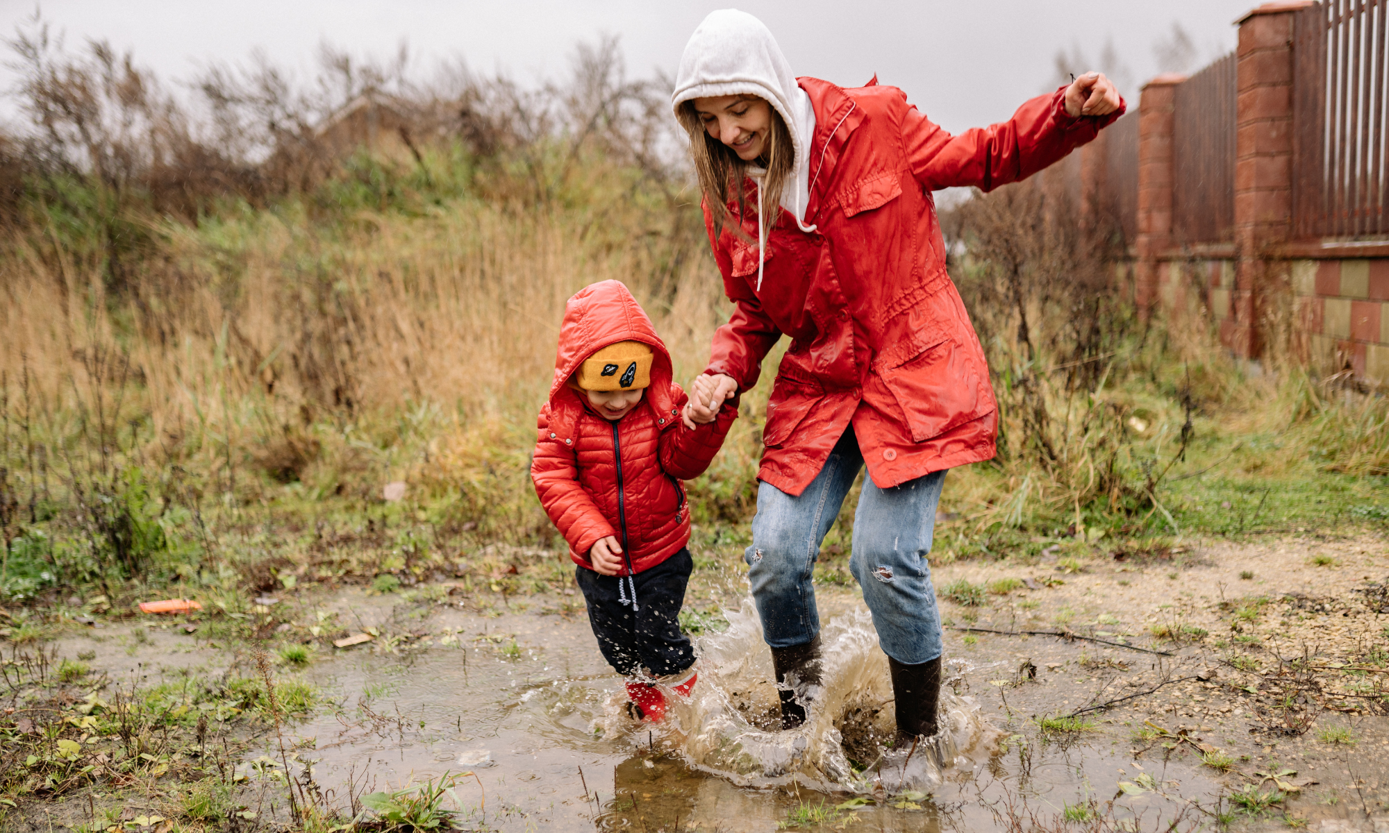 Mamma och barn i röda jackor hoppar glatt i en vattenpöl